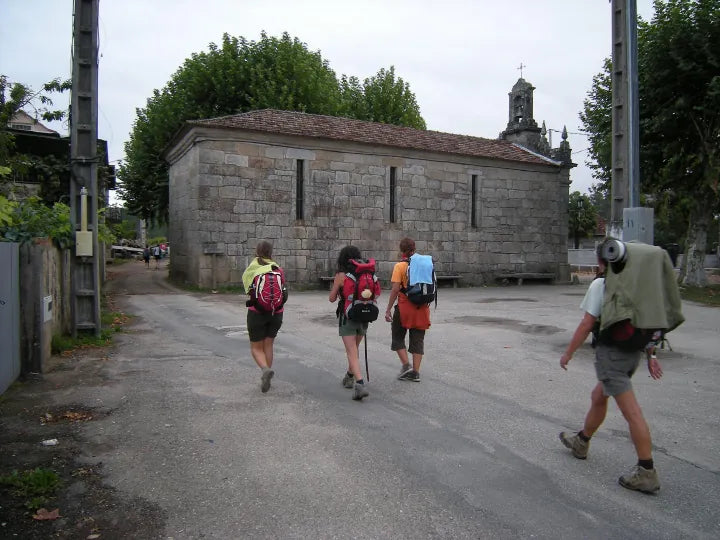 Cinco excursionistas con mochilas caminan por una calle asfaltada, pasando junto a una pequeña capilla de piedra. El día está nublado.
