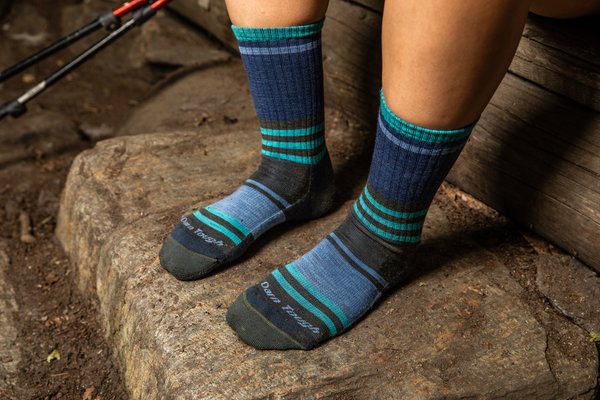 Dos piernas con calcetines multicolor azul, negro, púpura y turquesa sobre una piedra y al lado unos bastones.