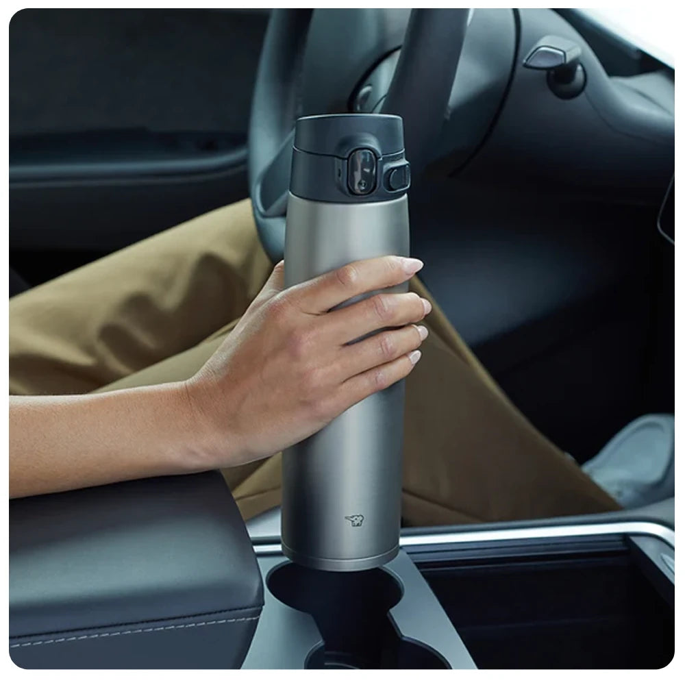 Termo Zojirushi SM-VA de acero inoxidable con tapa negra, sostenido por una mano en el portavasos de un automóvil. Se ve parte del volante y el interior del vehículo.