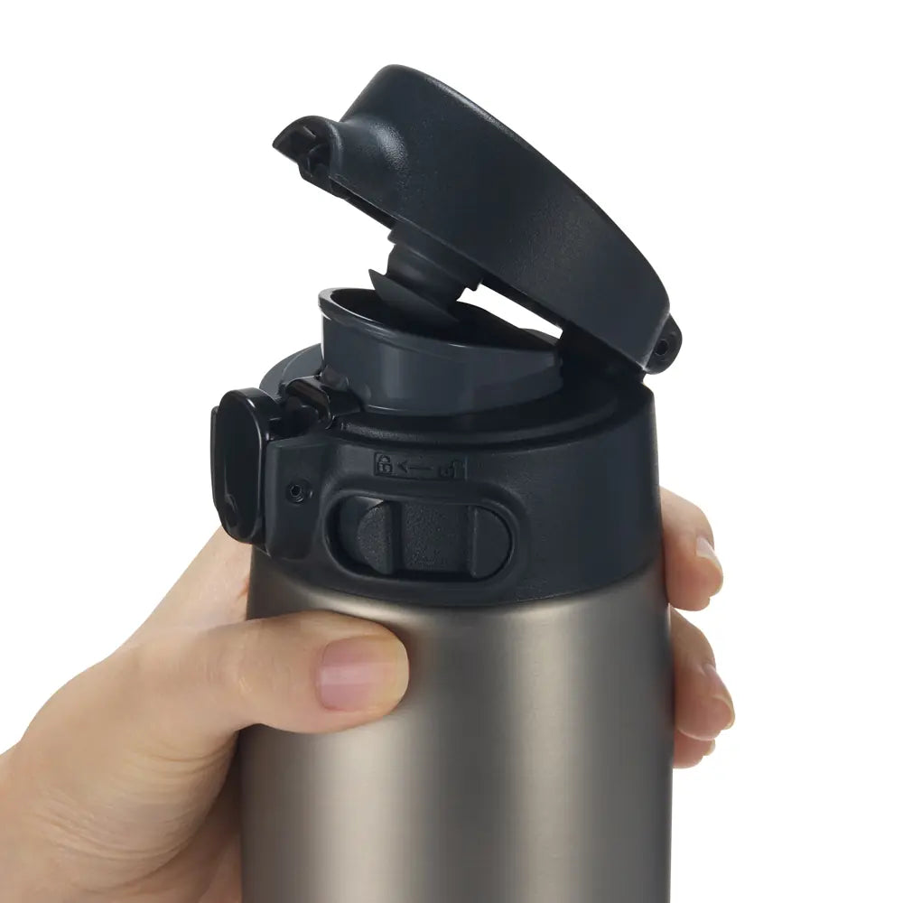 Mano sosteniendo una botella térmica Zojirushi plateada, mostrando en detalle la tapa negra abierta con su mecanismo de cierre y la boquilla para beber. Se aprecia el botón de bloqueo en la tapa y parte del cuerpo metálico de la botella.