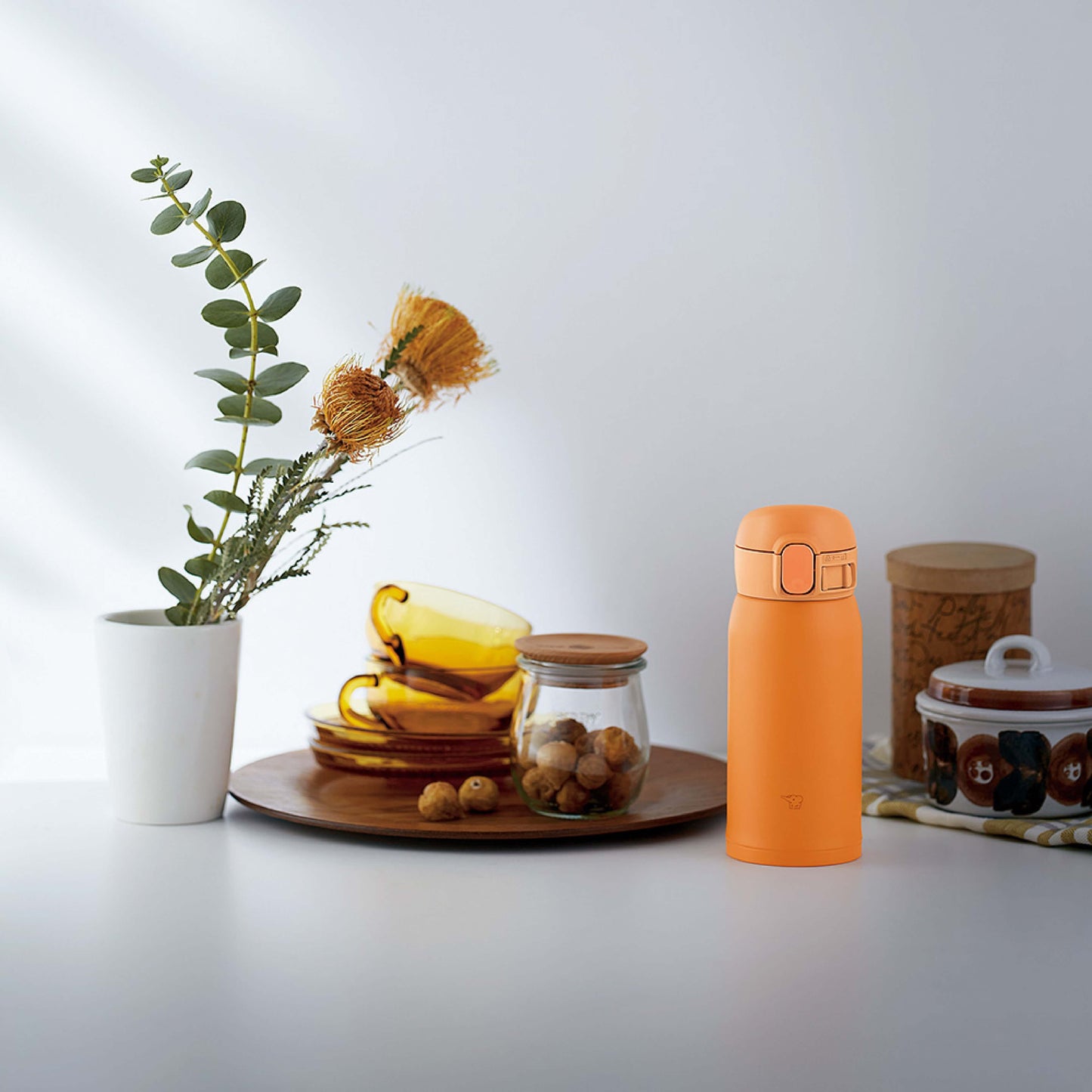 Escena de cocina con botella térmica Zojirushi SM-WA36 naranja junto a plato con tazas amarillas, frasco de nueces, planta en florero blanco y otros elementos decorativos sobre una superficie clara