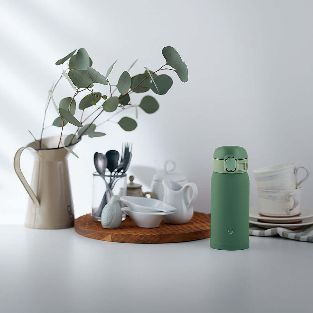 Escena de cocina con botella térmica Zojirushi SM-WA36 caqui junto a plato con tazas amarillas, frasco de nueces, planta en florero blanco y otros elementos decorativos sobre una superficie clara