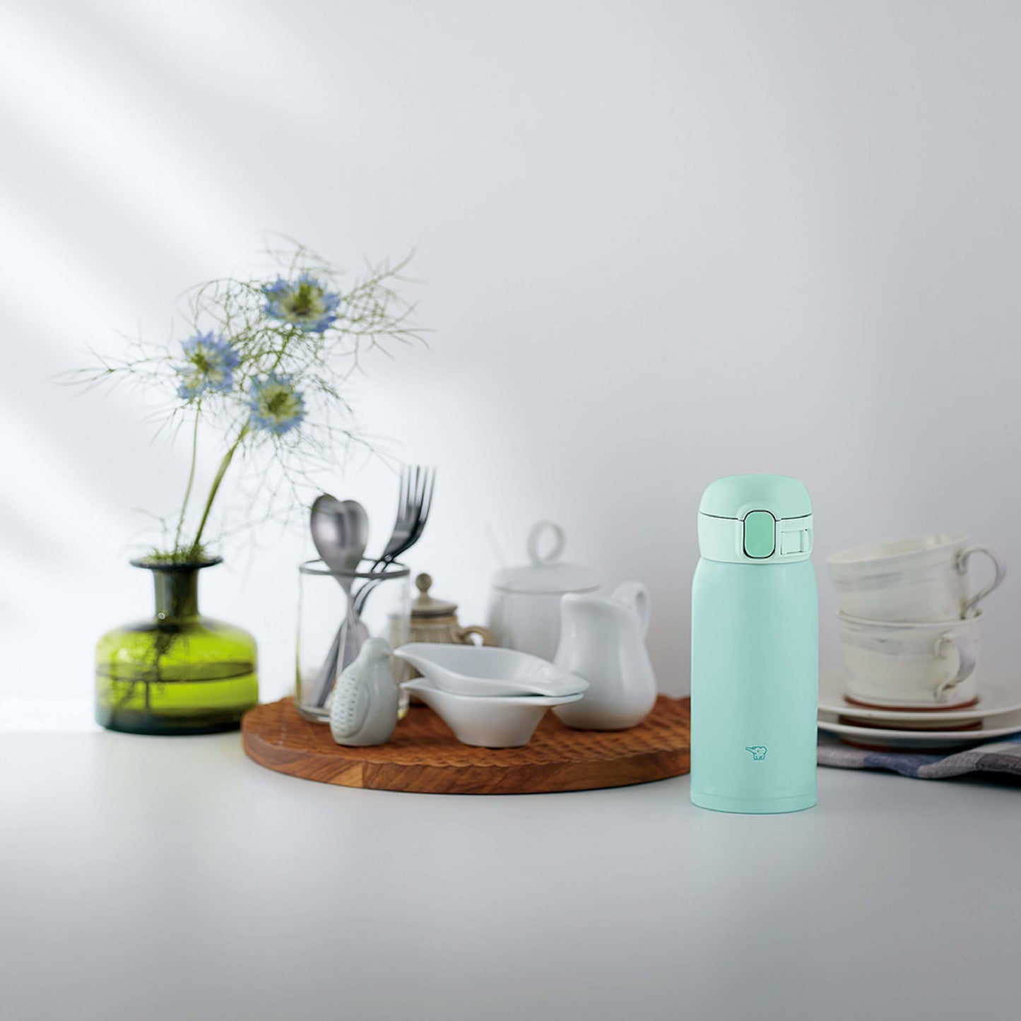Escena de cocina con botella térmica Zojirushi SM-WA36 verde junto a plato con tazas amarillas, frasco de nueces, planta en florero blanco y otros elementos decorativos sobre una superficie clara