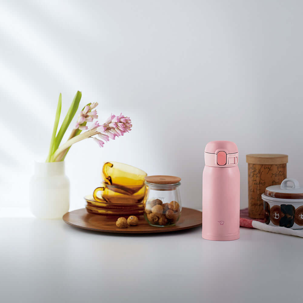 Escena de cocina con botella térmica Zojirushi SM-WA36 rosa junto a plato con tazas amarillas, frasco de nueces, planta en florero blanco y otros elementos decorativos sobre una superficie clara