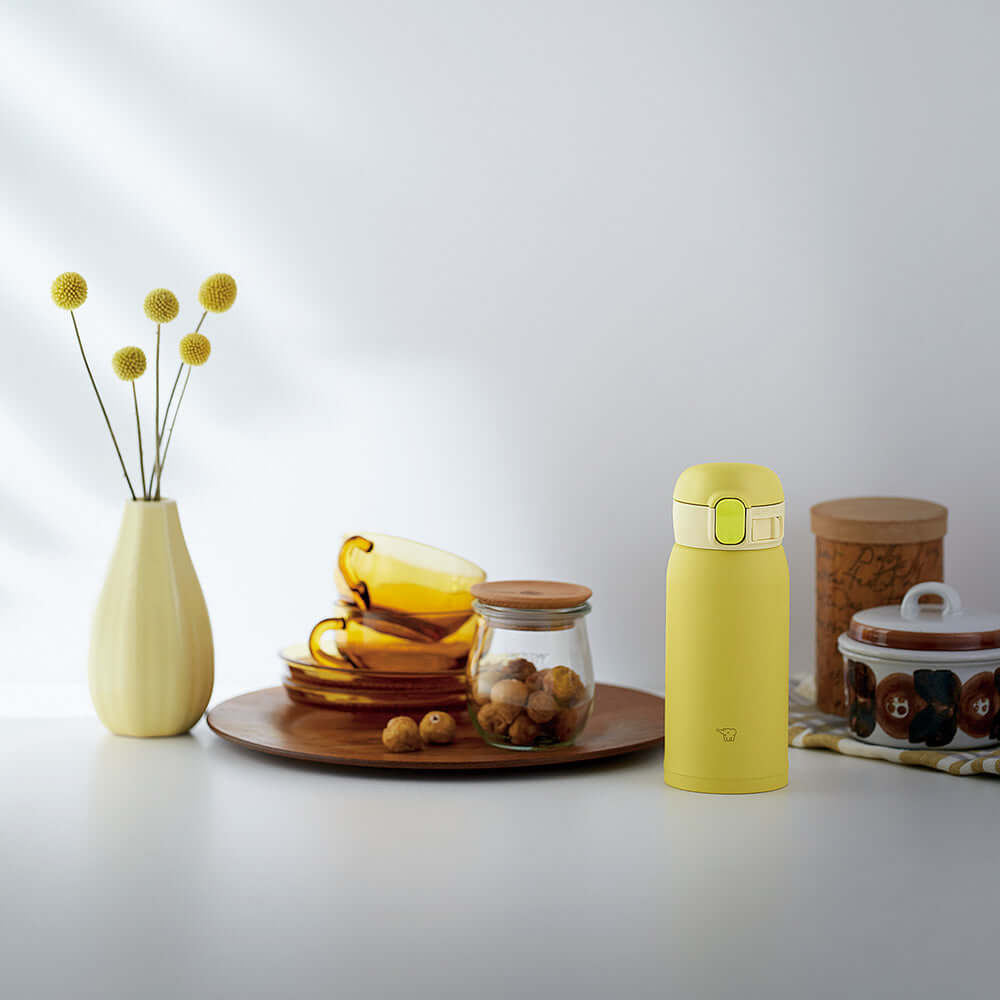 Escena de cocina con botella térmica Zojirushi SM-WA36 amarilla junto a plato con tazas amarillas, frasco de nueces, planta en florero blanco y otros elementos decorativos sobre una superficie clara