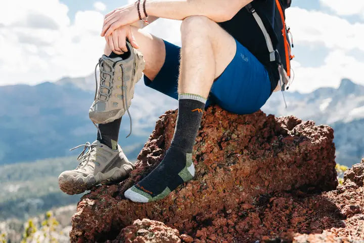 Persona sentada en una roca en la montaña, usando calcetines Darn Tough y sosteniendo una bota de senderismo en la mano.