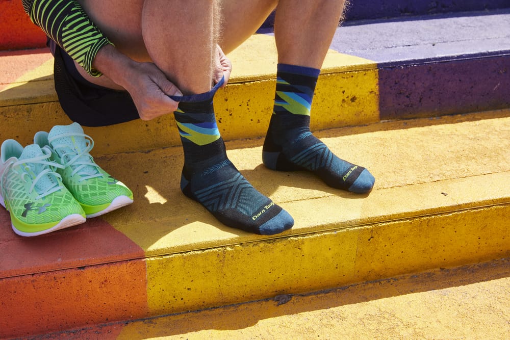 Persona poniéndose calcetines deportivos Darn Tough con colores vivos en una escalera de colores, junto a un par de zapatillas deportivas verdes