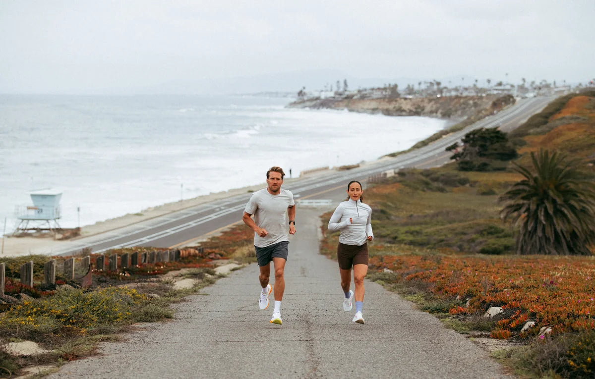 Dos personas corriendo por un camino junto a la playa, con el mar y colinas al fondo.