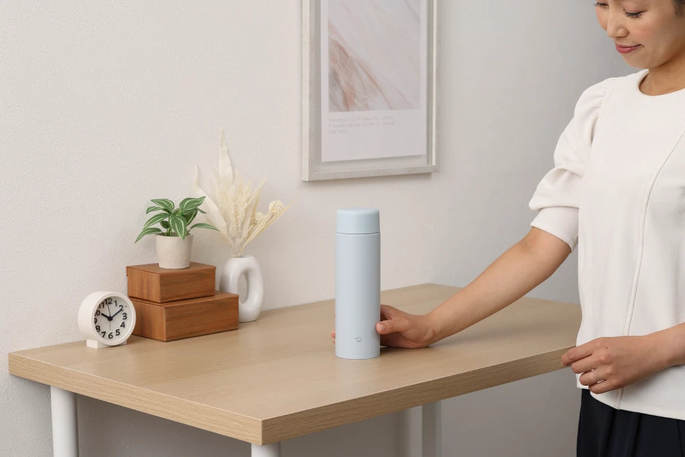 Alt text: Mujer tomando una botella térmica Zojirushi azul claro de una mesa decorada con elementos minimalistas y naturales