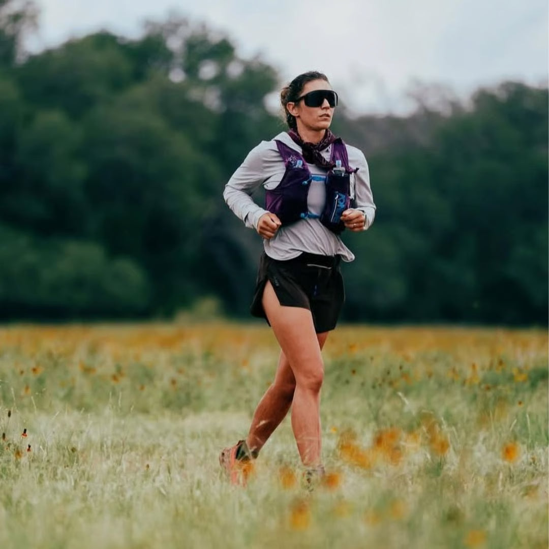 Mujer corriendo en un campo lleno de flores, lleva gafas de sol y un chaleco de hidratación.
