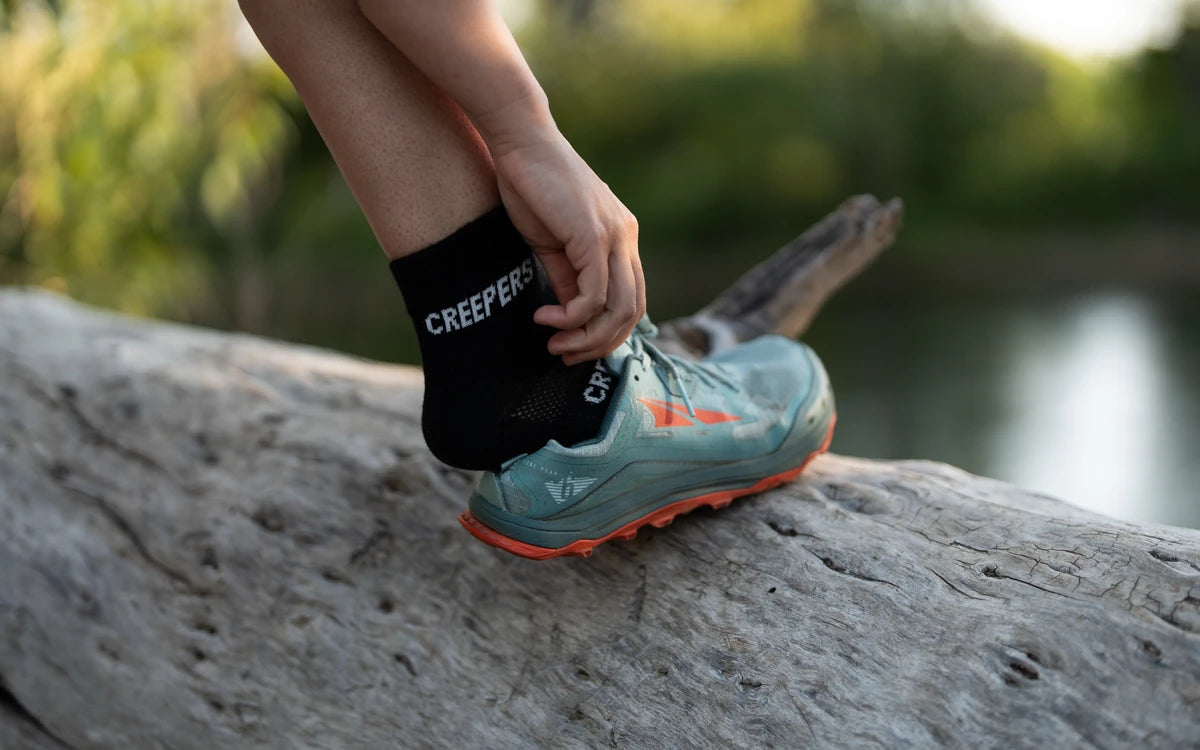Persona ajustando un calcetín negro con la palabra 'CREEPERS' mientras usa una zapatilla deportiva en un tronco.