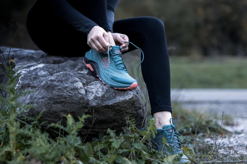 Persona atando los cordones de unas zapatillas deportivas azules sobre una roca, con fondo natural.