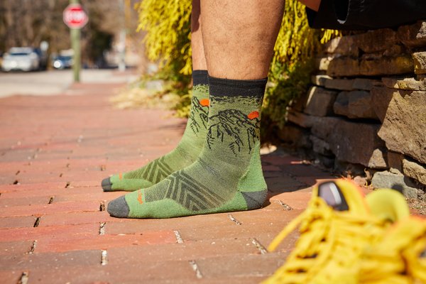 Dos piernas con calcetines verdes con la figura de una montaña en negro sobre un suelo color teja