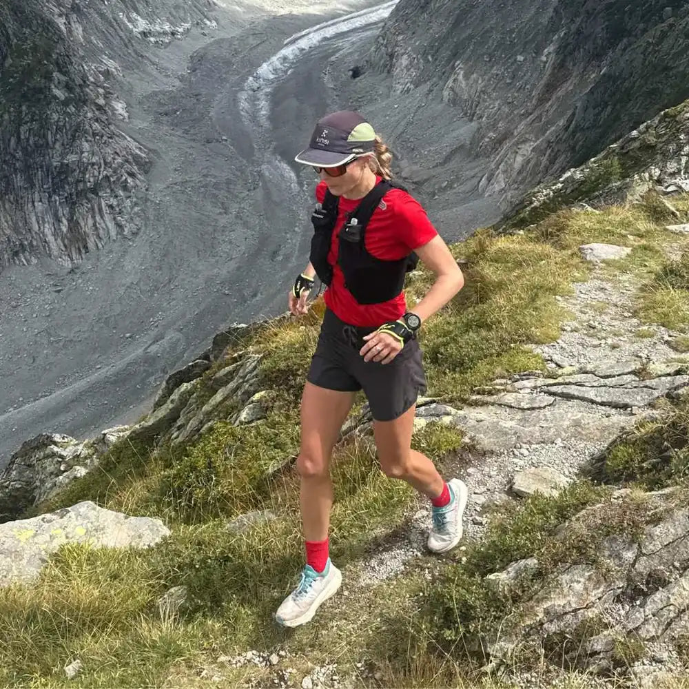 Corredora vestida con camiseta roja y pantalones cortos, corriendo por un sendero montañoso junto a un valle.