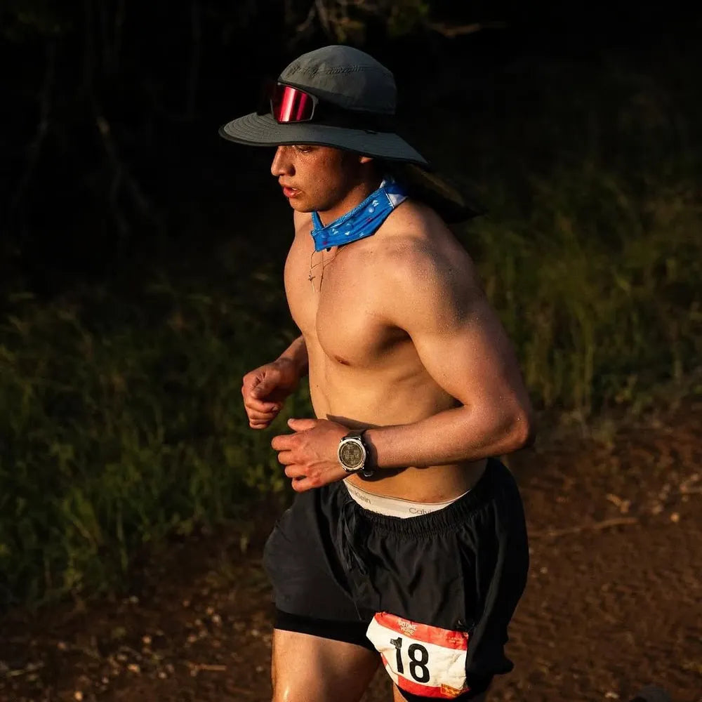 Corredor sin camiseta, usando un sombrero y gafas de sol, corre por un sendero al aire libre.