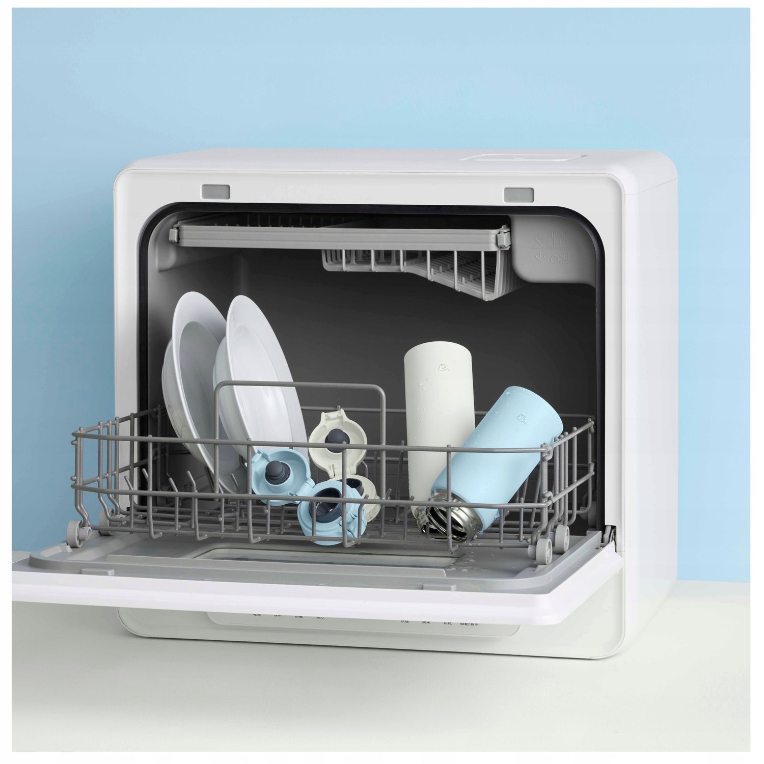 Fondo azul con lavavajillas blanco con platos blancos y una botella Zojirushi blanca y otra azul  con sus tapas.