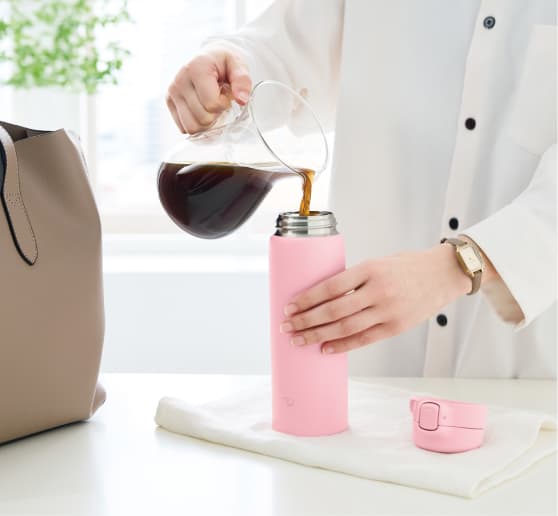 Modelo con camisa blanca y reloj dorado con un bolso marrón al lado vertiendo café con una jarra transparente en una botella rosa de Zojirushi.