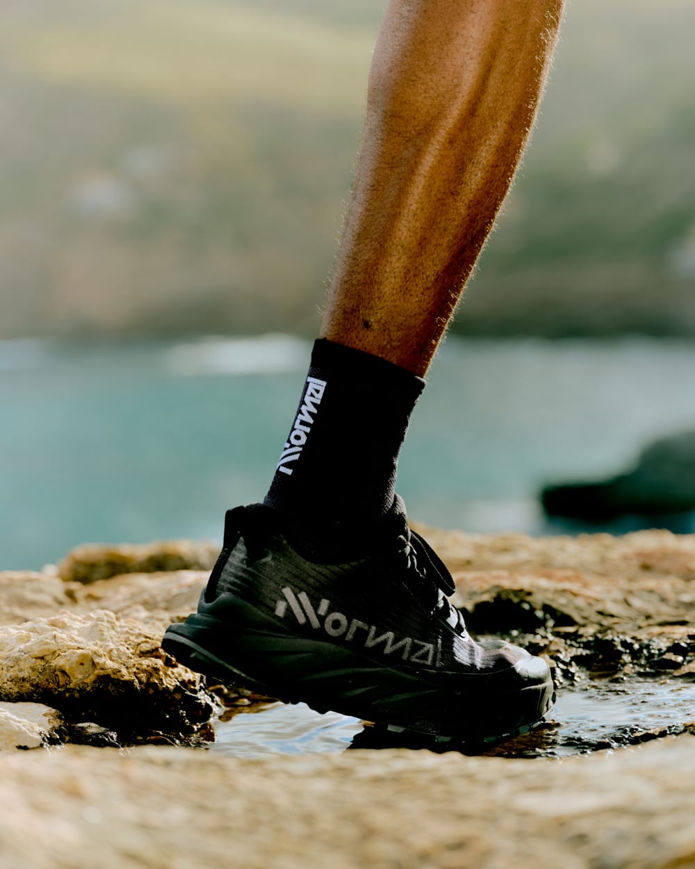 Un pie calzado con zapatillas negras sobre una roca, con agua alrededor y un paisaje natural de fondo.