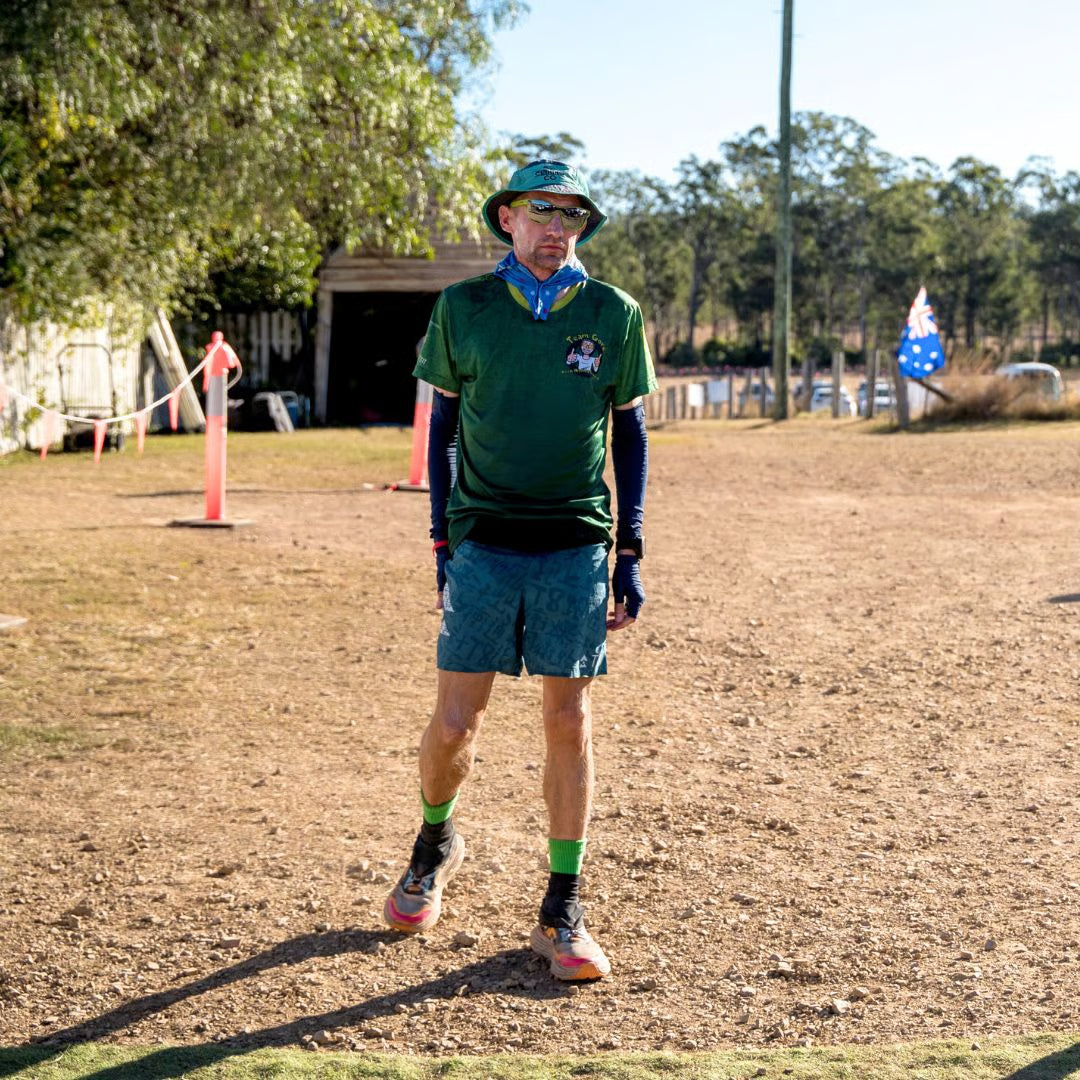 Hombre mayor vestido con camiseta verde y pantalones cortos, de pie en un terreno de tierra con árboles al fondo.