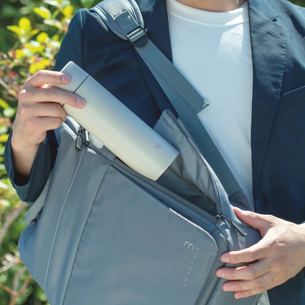 Persona con traje sosteniendo una botella térmica Zojirushi gris junto a un bolso de mensajero, en un entorno exterior con vegetación.