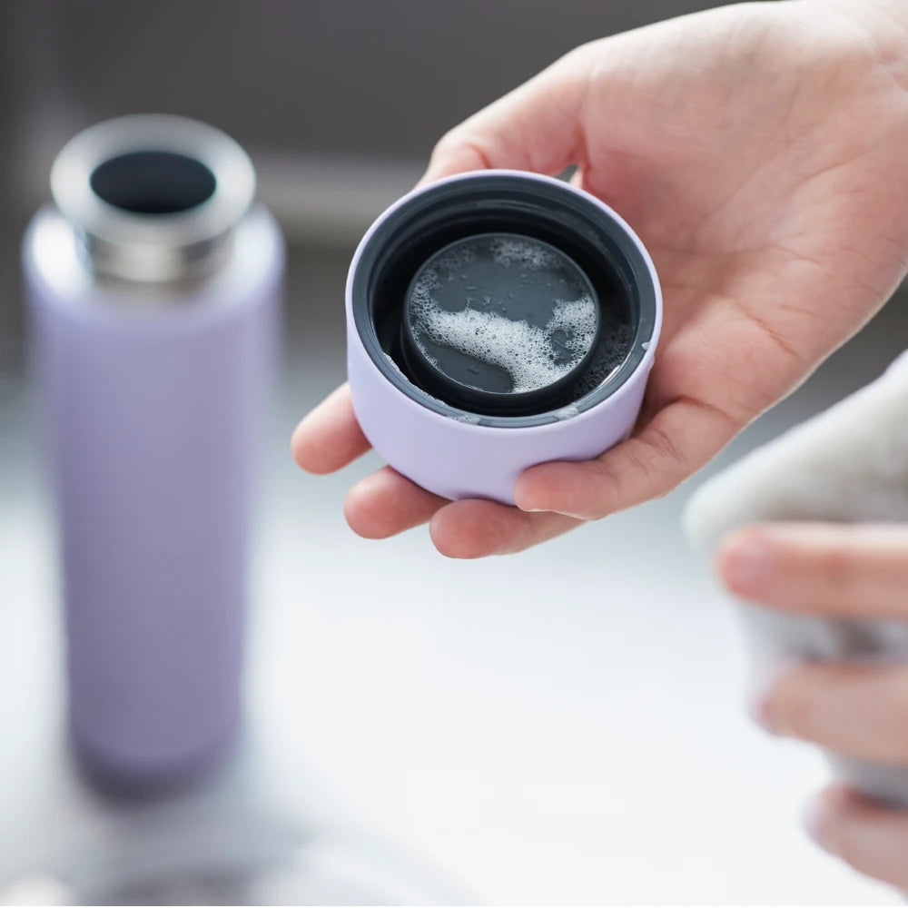 Mano sosteniendo la tapa de una botella térmica Zojirushi lila, con líquido visible en su interior. Botella completa de fondo