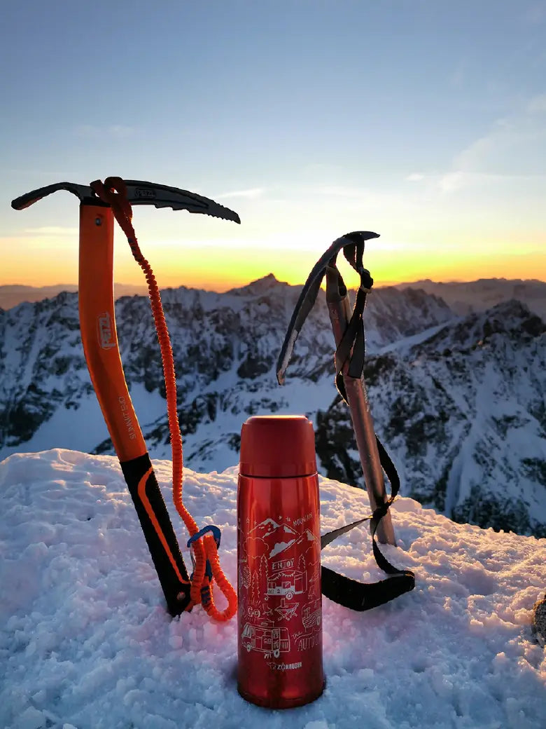 Pico de montaña al atardecer con dos piolets y un termo rojo en la nieve.