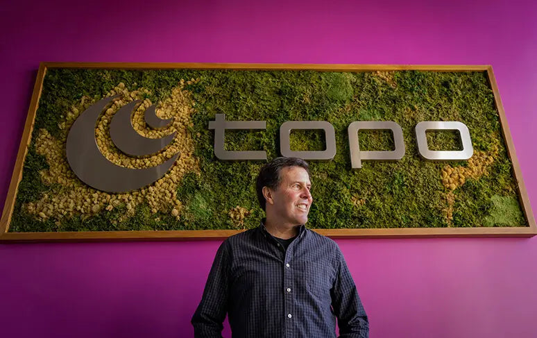 Fundador de Topo Athletic sobre un cuadro de hierba y tierra con logo y letras Topo en metal con fondo rosa