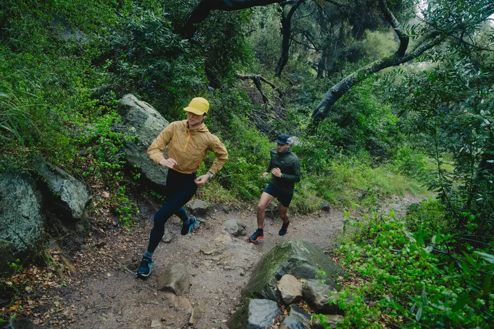Dos corredores en un sendero de montaña rodeados de vegetación y rocas, uno con gorra amarilla y otro con gorra oscura.